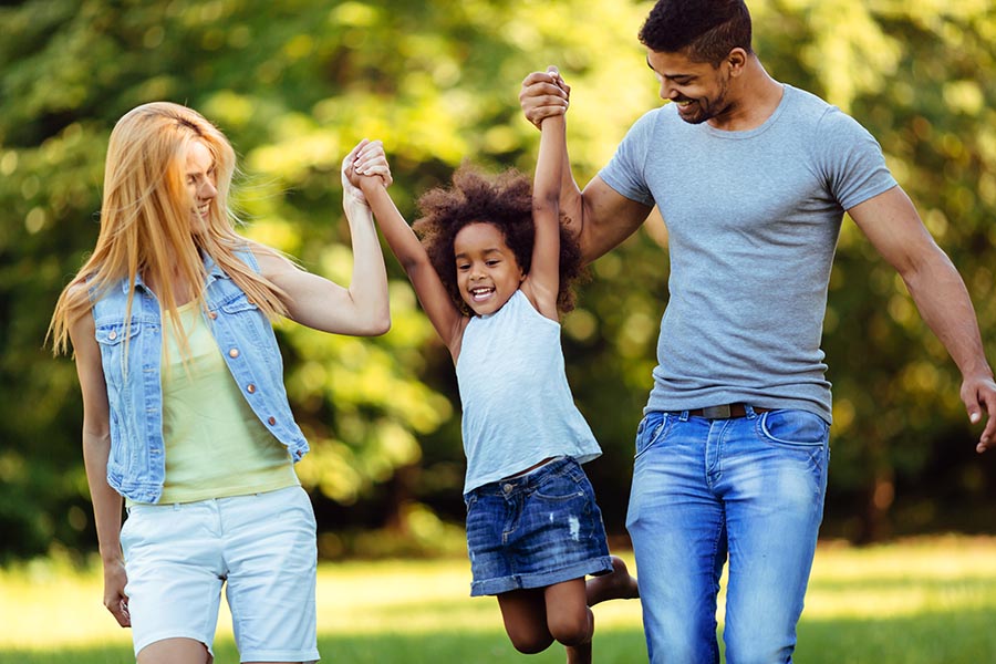 About Our Agency - Mother and Father Swing Their Daughter Into the Air Between Them, All Dressed for Warm Weather on a Sunny Day