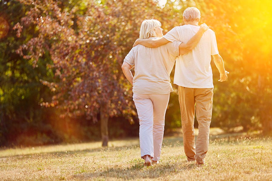Blog - Senior Couple Strolls Through Their Yard Arm-in-Arm, Sun Shining Through the Apple Trees