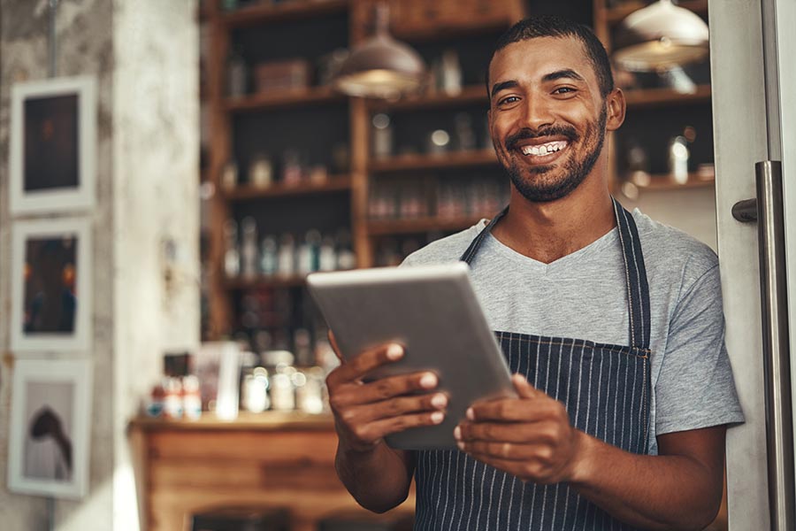 Business Insurance - Cafe Owner Stands in the Doorway of His Business Holding a Tablet, Smiling and Wearing an Apron